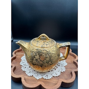 May include: A metallic gold teapot featuring a textured, mottled surface finish. The teapot has a rounded body, a short spout, a matching lid with a small handle, and a rectangular side handle. The gold finish has a reflective, uneven quality.