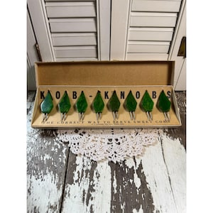 May include: A vintage set of eight green leaf-shaped corn holders in a cardboard box. The box reads "KAOBA-KANOBAS" and "THE CORRECT WAY TO SERVE SWEET CORN". The corn holders are green with metal prongs.