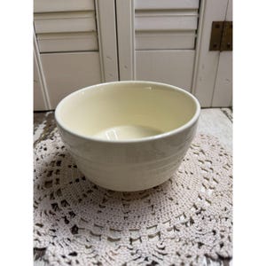Puede incluir: Un cuenco de cerámica de color crema con un acabado liso y brillante y tres ranuras horizontales que rodean el exterior. El cuenco presenta un borde circular ancho y abierto y una base profunda y redondeada, con un diseño limpio y sencillo.