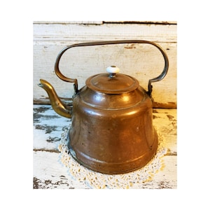 May include: A copper tea kettle with a wide, rounded body and a curved spout. The kettle features a tall, arched metal handle and a removable lid topped with a small, round white ceramic knob. The metal surface shows a warm, aged patina.
