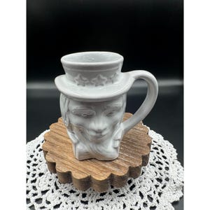 May include: A white ceramic mug shaped like a face wearing a top hat with star details. The mug has a handle and is sitting on a wooden coaster with a scalloped edge. The background is black.