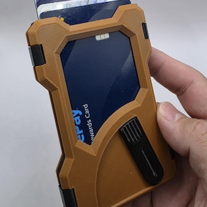 May include: A tan and black card holder wallet with a rigid, angular frame. The wallet features a central cutout displaying a blue rewards card with the visible text 'epay' and 'ewards Card', and includes a black clip attached to the front surface.