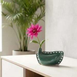Puede incluir: Un pequeño jarrón de cerámica verde oscuro con una forma única de barco, adornado con detalles de puntos blancos. Una vibrante flor rosa con centro amarillo se exhibe en el jarrón. El jarrón está sobre una superficie blanca.