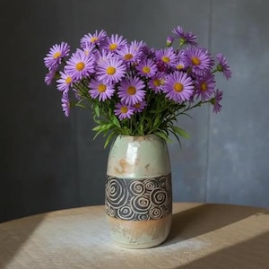 Puede incluir: Un ramo de flores de aster moradas con centros amarillos en un jarrón de cerámica. El jarrón tiene una parte superior verde claro, un diseño de remolino en blanco y negro en el medio y una base beige. El arreglo está sobre una mesa de madera.
