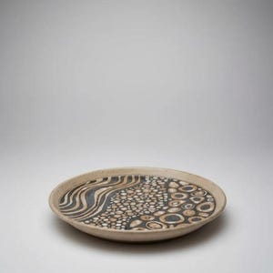 Puede incluir: Un plato de cerámica hecho a mano con un borde beige y un interior estampado. El diseño presenta una sección ondulada a rayas negras y beige, una sección de pequeños círculos beige y círculos negros y beige.