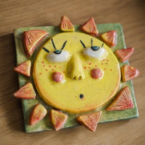 May include: A ceramic sun face with a yellow face, red freckles, and orange sun rays. The sun is set on a green square base. The sun's eyes are blue and the cheeks are pink. The sun has a surprised expression.