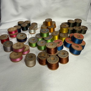 May include: A collection of vintage wooden spools of thread in various colors, including red, pink, blue, green, yellow, and brown. The spools are arranged on a white surface, showcasing the variety of thread colors and the aged appearance of the wooden spools.