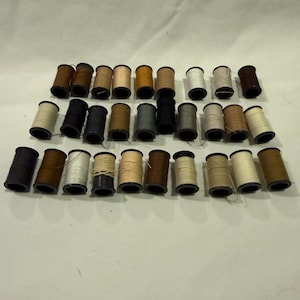 May include: A collection of twenty-six sewing thread spools arranged in three rows. The spools feature various neutral colors including shades of brown, tan, beige, gray, black, and white, with visible thread wound around each cylindrical core.