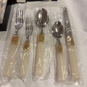 May include: A set of five pieces of silverware, including forks, spoons, and a knife, each wrapped in plastic. The silverware features ivory-colored handles with gold-colored accents.
