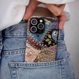 May include: A decorative phone case with a patchwork design featuring floral and bird motifs in shades of blue, brown, and pink. The case is partially visible in the back pocket of a pair of blue jeans.