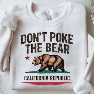 May include: White sweatshirt with the text "DON'T POKE THE BEAR" in brown letters. Below the text is a brown bear graphic, a red star, and the words "CALIFORNIA REPUBLIC". A red stripe is at the bottom.