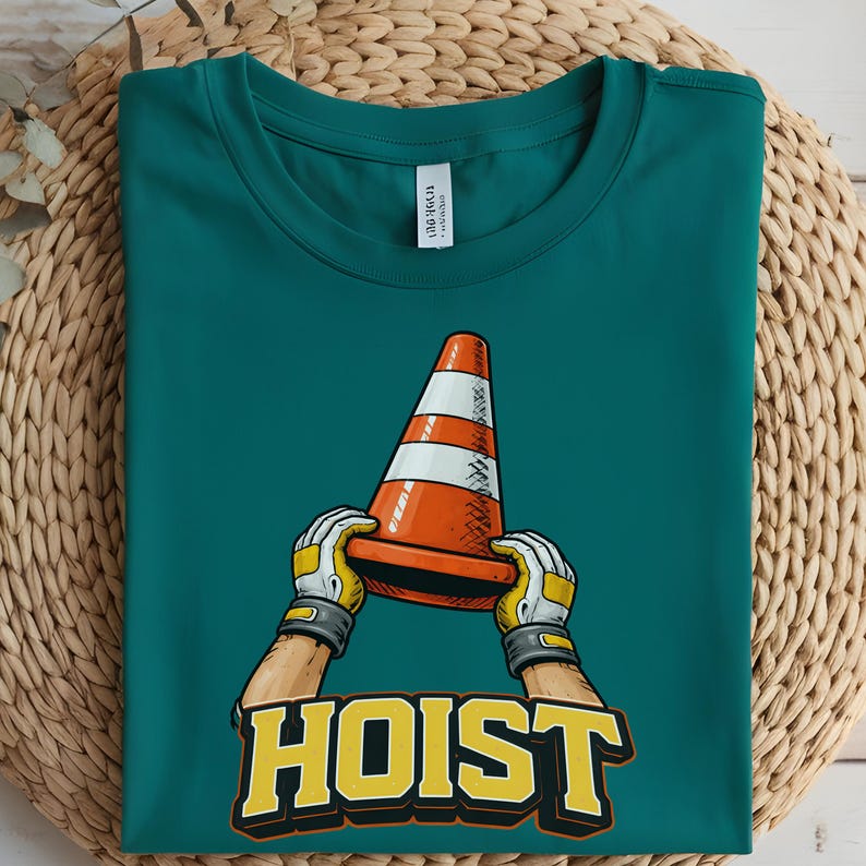 May include: Teal t-shirt featuring a graphic of a traffic cone held by gloved hands, with the word "HOIST" in yellow and black block letters. The shirt is folded on a woven surface.