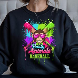 May include: Black sweatshirt featuring a cartoon monkey wearing pink sunglasses and a bow tie, with crossed baseball bats. The design includes the text "Party Animals Baseball" in a colorful, dripping paint style.