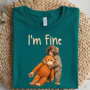 May include: Teal t-shirt with the text "I'm Fine" in a light beige color. The shirt features a graphic of two monkeys hugging. One monkey is brown and the other is orange.
