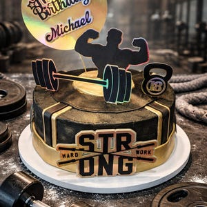 May include: A black and gold birthday cake with a fitness theme. The cake is decorated with a barbell, a weight, and a silhouette of a bodybuilder. The cake has the words "Happy Birthday Michael" and "Strong Hard Work".