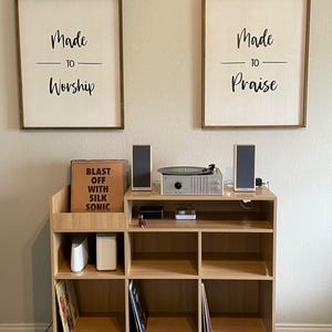 May include: A wooden record player stand with a built-in record storage compartment. The stand has a turntable, speakers, and two framed signs that read "Made to Worship" and "Made to Praise". The record stand is made of light-colored wood.
