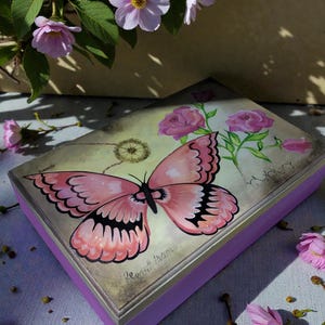 Puede incluir: Una caja rectangular de madera con base lila y tapa plateada. La tapa está decorada con una gran mariposa rosa y rosas rosas. La caja está rodeada de flores rosas.