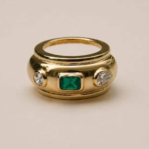 Puede incluir: Un anillo de tono dorado con una piedra preciosa rectangular verde en el centro, flanqueada por dos piedras transparentes facetadas. La banda es ancha y lisa con un acabado pulido, mostrando una disposición simétrica de las tres piedras.
