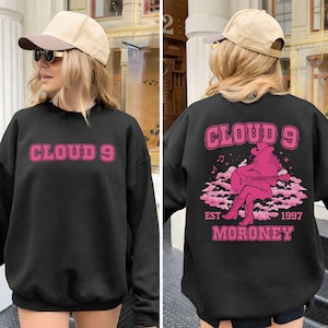 Puede incluir: Sudadera negra con el texto "CLOUD 9" en rosa en la parte delantera. La parte trasera presenta un gr&aacute;fico rosa de una vaquera tocando la guitarra en una nube, con el texto "CLOUD 9 EST 1997 MORONEY".