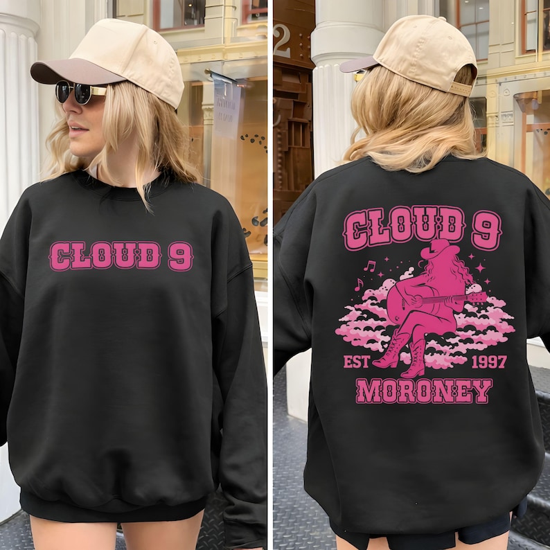 Puede incluir: Sudadera negra con el texto "CLOUD 9" en rosa en la parte delantera. La parte trasera presenta un gr&aacute;fico rosa de una vaquera tocando la guitarra en una nube, con el texto "CLOUD 9 EST 1997 MORONEY".