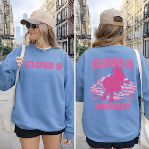 Puede incluir: Una sudadera azul claro con el texto "CLOUD 9" en letras rosas en la parte delantera. La parte trasera presenta un gr&aacute;fico rosa de una persona tocando la guitarra sobre nubes, con el texto "EST 1997 MORONEY".