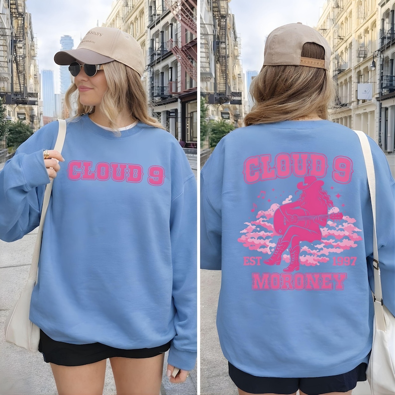Puede incluir: Una sudadera azul claro con el texto "CLOUD 9" en letras rosas en la parte delantera. La parte trasera presenta un gr&aacute;fico rosa de una persona tocando la guitarra sobre nubes, con el texto "EST 1997 MORONEY".