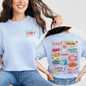 May include: Light blue t-shirt with the text "CLOUD 9" on the front. The back of the shirt features various phrases and words in colorful text boxes, including "Stupid," "Medicine," and "Beautiful Things."