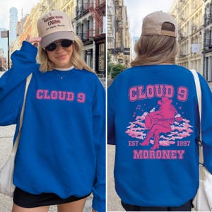 Puede incluir: Sudadera azul real con el texto "CLOUD 9" en rosa en el pecho. La parte trasera presenta un gr&aacute;fico rosa de una vaquera tocando la guitarra sobre nubes, con "EST 1997 MORONEY" debajo. Se usa una gorra color canela.