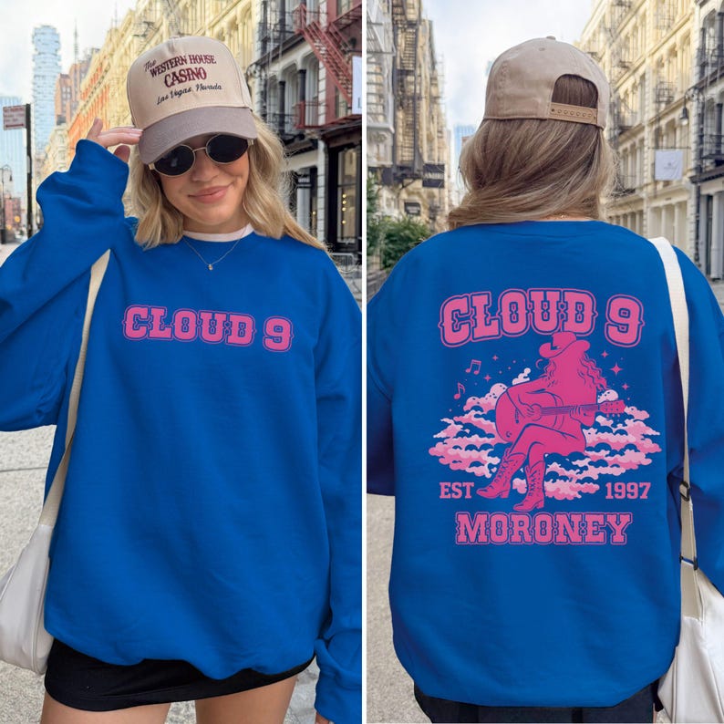 Puede incluir: Sudadera azul real con el texto "CLOUD 9" en rosa en el pecho. La parte trasera presenta un gr&aacute;fico rosa de una vaquera tocando la guitarra sobre nubes, con "EST 1997 MORONEY" debajo. Se usa una gorra color canela.