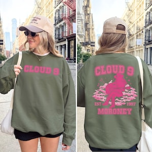 Puede incluir: Sudadera verde oliva con el texto rosa "CLOUD 9" en la parte delantera. La parte trasera presenta un gr&aacute;fico rosa de una persona sentada en las nubes, con "CLOUD 9 EST 1997 MORONEY". Sudadera de estilo casual.