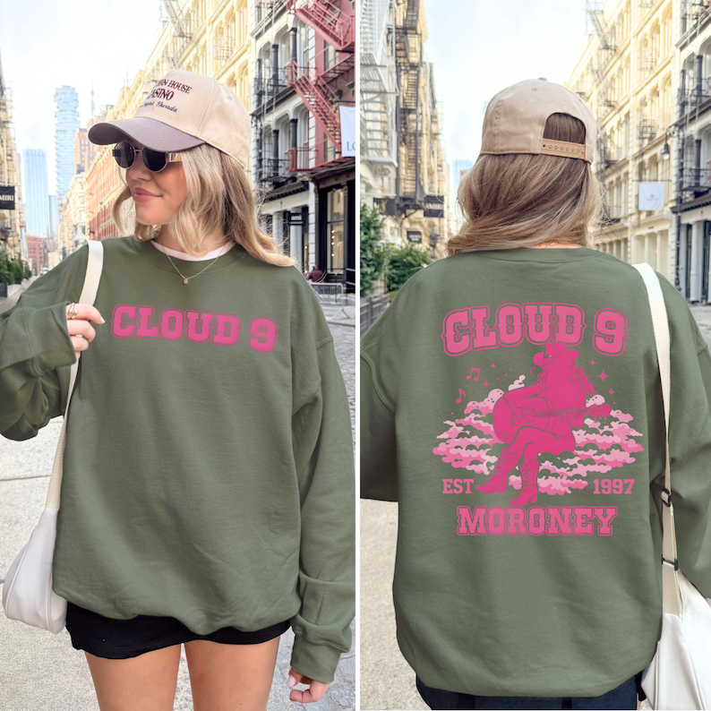 Puede incluir: Sudadera verde oliva con el texto rosa "CLOUD 9" en la parte delantera. La parte trasera presenta un gr&aacute;fico rosa de una persona sentada en las nubes, con "CLOUD 9 EST 1997 MORONEY". Sudadera de estilo casual.