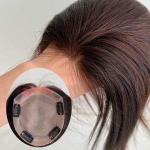 Op de afbeelding: Een donkerbruine haartop met een kanten basis en clips voor bevestiging. Het haarstuk is ontworpen om volume en dekking toe te voegen. Het haar is gestyled met een natuurlijke scheiding en lijkt mensenhaar te zijn.