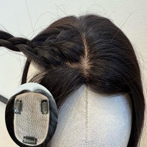 Pode incluir: Um topper de cabelo castanho escuro com uma seção trançada. A peça capilar apresenta uma base de renda com três clipes para fixação segura. O cabelo é estilizado para mostrar a divisão e uma linha de cabelo de aparência natural.