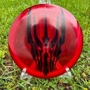 Può includere: Un disco da disc golf rosso con un design grafico nero. Il disco è esposto su un supporto di plastica trasparente, su uno sfondo di erba verde. La grafica è un design stilizzato e simmetrico.
