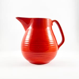 May include: A bright red ceramic pitcher featuring a rounded, bulbous body with horizontal ribbed detailing throughout. The vessel includes a prominent pouring spout on the left side and a curved handle on the right side, finished in a glossy glaze.