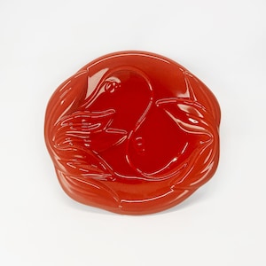 May include: A circular, glossy red ceramic dish featuring a raised, embossed design of two stylized fish swimming in a circular pattern, creating a yin-yang effect. The surface is smooth and reflective with deep red tones throughout the piece.