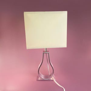 May include: A table lamp featuring a square, off-white fabric lampshade atop a silver-toned metal base. The base has a hollow, teardrop-shaped frame that connects to a rectangular pedestal. A white power cord extends from the base of the lamp.