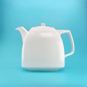 May include: A white ceramic teapot with a square-shaped body, a short spout, a flat lid with a small knob, and a wide angular handle. The teapot features smooth, clean lines and a glossy finish, presented against a solid light blue background.