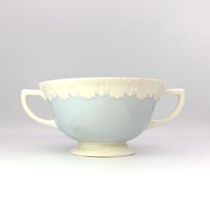 May include: A two-handled ceramic bowl with a light blue body and a cream-colored rim. The bowl has a pedestal base and two curved handles. The rim has a textured, uneven edge. The bowl is likely for serving soup or other dishes.