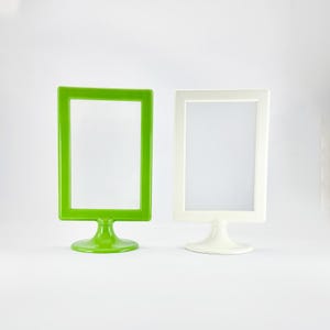 May include: Two vertical rectangular sign holders with a smooth finish, one in bright lime green and one in solid white. Each frame is supported by a circular pedestal base, designed to display signage or menus in an upright, stable position.