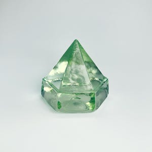 May include: A translucent light green glass paperweight shaped like a faceted pyramid with a hexagonal base. The object features smooth, polished surfaces that catch the light, creating subtle reflections and internal depth within the glass.