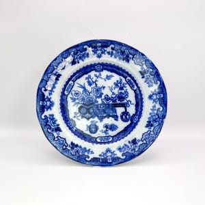 May include: A round, white plate with a blue floral design. The plate features a central scene with a table, flowers, and vases, surrounded by a detailed border. The plate's edge has a repeating floral pattern.
