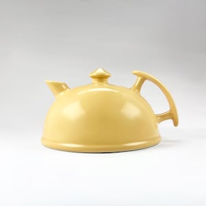 May include: A pale yellow ceramic teapot with a smooth, glossy finish. The teapot features a low, rounded dome-shaped body, a short spout, a small central lid knob, and a curved handle that extends from the side to the top of the vessel.