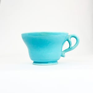May include: A bright turquoise ceramic teacup with a smooth, glossy finish. The cup features a wide, flared rim, a rounded body, and a distinct pedestal base. A curved, ornate handle is attached to the side, completing the solid color design.