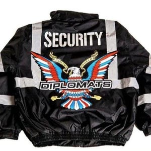 Pode incluir: Casaco de segurança preto com faixas refletoras e a palavra "SECURITY" em letras brancas nas costas. Apresenta um emblema de águia colorido com as palavras "HARLEM WORLD DIPLOMATS".
