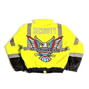Pode incluir: Casaco de segurança amarelo brilhante com faixas refletoras prateadas e mangas pretas. A parte de trás apresenta um emblema de águia colorido com a palavra "DIPLOMATS" e a palavra "SECURITY" no topo.