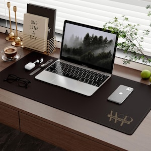 May include: A dark brown desk mat with a laptop, smartphone, and accessories. The laptop displays a forest scene. Other items include a pair of glasses, wireless earbuds, and a watch. The mat has the text "WARNER & PENCER".