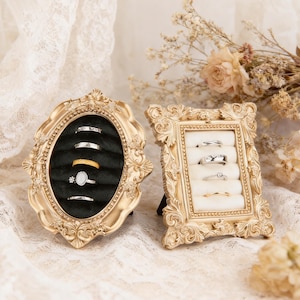 May include: Two ornate gold-colored ring holders. One is oval with a black velvet interior, displaying rings. The other is rectangular with a cream-colored interior, also displaying rings. Both have decorative frames.