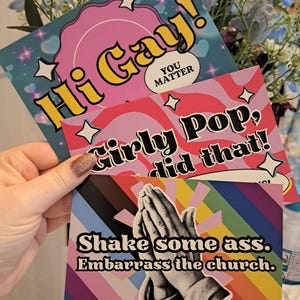 May include: Three vibrant greeting cards with striking text and designs. One card says "Hi Gay!" with a pink circle and stars. Another reads "Girly Pop, did that!" with a wavy pink pattern. The third card shows praying hands and the text "Shake some ass. Embarrass the church."