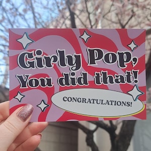 May include: A greeting card with the text "Girly Pop, You did that!" in a retro font. The card has a pink and red wavy background with star accents and the word "Congratulations!" in a white oval.
